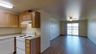 This single-floor space offers an open-concept layout that seamlessly connects the kitchen and living area, creating a cohesive and functional living environment. The kitchen is equipped with warm-toned wood cabinetry featuring ample upper and lower storage, a white electric coil range with an overhead ventilation hood, and light-colored countertops. The under-cabinet lighting adds a practical touch to the workspace.

The flooring throughout the main living area consists of dark hardwood-style planks that provide a clean, modern contrast to the neutral beige walls. A ceiling fan with a light fixture is centrally positioned in the living area, contributing to both air circulation and ambient lighting.

At the far end of the living space, a large sliding glass door floods the interior with natural light and opens to what appears to be a private balcony enclosed by a railing, offering an outdoor extension of the living space. The open sightlines from the kitchen through to the living area enhance the sense of spaciousness, making the unit feel bright and airy despite its compact footprint.