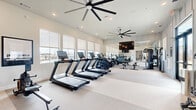 Located in Hutto, TX, this well-equipped fitness center offers a bright, open layout purpose-built for a complete workout experience. The space is finished with light neutral tones, recessed lighting, and large windows fitted with roller shades that allow natural light to filter in while framing views of an outdoor pool area. Oversized ceiling fans provide airflow throughout, enhancing comfort during workouts.

The cardio zone is prominently featured along the windowed wall, housing a row of TRUE-brand treadmills and elliptical machines, each equipped with individual digital display screens. Adjacent to the cardio equipment, a multi-station cable machine and a set of weight benches provide dedicated strength training options. A dumbbell rack stocked with free weights rounds out the strength area near the entrance.

Full-length mirrors line one wall, amplifying the sense of space and allowing users to monitor their form. A wall-mounted flat-screen television is centrally positioned for viewing during workouts. The flooring consists of a light-toned carpet that softens the space while maintaining a clean, modern aesthetic. Glass entry doors with black frames contribute to the contemporary design. The overall ambiance is clean, functional, and welcoming, making it a well-appointed facility for residents of the Hutto, TX community.