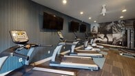 This modern fitness center is thoughtfully designed to support a comprehensive workout experience. The space is organized into distinct functional zones, seamlessly flowing from cardio to strength training areas. A row of True-brand treadmills and elliptical machines lines one side of the room, each positioned in front of wall-mounted flat-screen televisions — ideal for staying entertained during longer cardio sessions. The accent wall behind the cardio equipment features a bold, textured blue-gray paneling that adds a sleek, contemporary character to the space. On the opposite end, a large-scale black-and-white photographic mural depicting dynamic athletic imagery — including cycling, running, and yoga — serves as a striking and motivating focal point. Adjacent to the mural, a cable strength machine and a weight bench provide dedicated resistance training options, complemented by a dumbbell rack for free weight exercises. The flooring consists of a patterned carpet tile in charcoal and teal, adding visual interest while providing comfort underfoot. A full-length mirror expands the sense of space and supports form-checking during workouts. Multiple windows along one wall allow natural light to flow in, offering views of the outdoors and creating an open, energizing atmosphere. Recessed lighting throughout ensures the space is well-lit at all times.