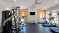 This fitness center is a spacious, single-floor facility designed to accommodate a comprehensive workout experience. The space features vaulted ceilings accented by recessed lighting and ceiling fans, contributing to an open, airy atmosphere. Warm amber accent walls contrast with crisp white surfaces, creating an energizing environment for exercise.

The cardio section is well-stocked with multiple treadmills, elliptical trainers, and a stationary bike, each positioned near large windows that provide views of the surrounding greenery and abundant natural light. Wall-mounted flat-screen televisions are stationed throughout the space, keeping members entertained during workouts.

The strength training area includes a multi-station cable machine, a power tower, and a dumbbell rack with a range of free weights. A rowing machine and adjustable bench add further versatility to the equipment offering. A full-length wall mirror expands the visual depth of the space while allowing users to monitor their form.

Dark rubber flooring spans the entire facility, providing a durable, cushioned surface suitable for all types of exercise. Large multi-pane windows along multiple walls ensure consistent natural light throughout the day. Glass entry doors provide a welcoming entry point, enhancing the overall accessibility and open feel of the fitness center.