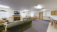 Located at 307 E Seventh St in Monticello, Minnesota, this single-floor common area space offers a comfortable and functional environment designed for communal gathering. The layout flows naturally from a welcoming entryway — accented by decorative wall art and a convenient console table — into a generously sized open living and dining area.

The lounge area is a central highlight, featuring multiple plush green sofas and armchairs arranged around a warm fireplace topped with a flat-screen television. Natural light filters in through several windows, and a decorative wallpaper border runs throughout the space, lending a cohesive and homey aesthetic. Textured ceilings and flush-mounted fluorescent lighting provide a bright, even illumination throughout.

The dining area accommodates a large wooden table with chairs and benefits from a sliding glass patio door that opens to an exterior view, further enhancing the sense of openness and connectivity to the outdoors. A wall-mounted air conditioning unit ensures year-round comfort.

The fully equipped kitchen features warm oak cabinetry, ample countertop space, a full suite of white appliances including a range, microwave, dishwasher, and refrigerator, as well as a central island with barstool seating and easy-clean tile flooring. The space is practical, well-organized, and suited for group gatherings or events.