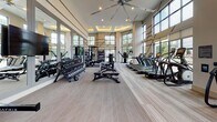 This single-floor fitness center is designed to offer a premium workout experience, combining thoughtful architecture with a full suite of modern equipment. The space is defined by soaring double-height ceilings and expansive floor-to-ceiling windows that flood the interior with natural light and offer open views of the surrounding outdoors, creating an energizing and airy atmosphere. A large ceiling fan with a coffered, softly lit surround adds both visual interest and comfort to the main workout area.

The flooring throughout features a neutral, patterned carpet that delineates the space cleanly. Along one side, a well-stocked dumbbell rack and free weight area is complemented by full-length mirrors that enhance the sense of space. A Matrix power rack with weight plates and benches provides a dedicated strength training zone. Cardio enthusiasts are well served by a row of treadmills, elliptical trainers, stair climbers, and stationary cycling bikes positioned near the windows.

A functional trainer cable machine occupies a prominent position near the entrance area, which transitions into a welcoming reception zone featuring hardwood flooring, a cubby storage unit, a water station, and contemporary wall art. The overall design strikes a balance between functionality and refined aesthetics, making this fitness center both practical and visually appealing.