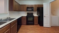 This single-story 1-bedroom unit in Lawton, Oklahoma offers a practical and comfortable layout designed for easy everyday living. The living area features plush carpeting and crown molding, with a front entry door and an additional door providing access to the outdoors, allowing natural light to fill the space. A pass-through breakfast bar connects the living area to the kitchen, creating an open and connected feel. The kitchen is well-equipped with rich wood cabinetry, light countertops, and a full suite of black appliances including a refrigerator, electric range, microwave, and dishwasher, all set atop warm wood-style flooring. The bedroom is generously sized with soft carpeting, dual windows that invite in natural light, and a walk-in closet with built-in shelving for ample storage. The full bathroom is cleanly finished with wood-style flooring, a combined tub and shower, a toilet, and a vanity with dark wood cabinetry and overhead storage. Throughout the unit, neutral wall tones and consistent finishes create a cohesive and move-in ready atmosphere.