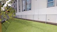 Located at 555 N Spring St in the heart of Downtown Los Angeles, this ground-level outdoor space serves as a dedicated pet area for the building's residents. The space is thoughtfully divided into two distinct zones: a lush artificial turf lawn, ideal for pets to lounge and play, and a sandy, open run area that provides a natural surface for more active use. Both zones are fully enclosed by sleek metal fencing, ensuring a safe and secure environment for pets and their owners alike.

A standout feature of the space is a vivid mural adorning the building's exterior wall, depicting silhouetted figures in teal, purple, and blue tones that add a lively, artistic character to the area. Raised planter beds with ornamental grasses and native plantings contribute additional greenery and texture throughout.

Pet waste stations are conveniently positioned within the space, promoting cleanliness and ease of use. The surrounding urban context is visible beyond the perimeter walls, offering views toward iconic Downtown LA landmarks, including City Hall and palm tree-lined boulevards, with LA Phil banners visible in the distance. The open-air design allows for ample natural light and fresh air, making this a functional and well-appointed amenity space in one of Los Angeles' most vibrant neighborhoods.