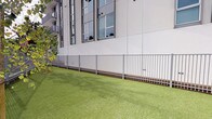 Located at 555 N Spring St in the heart of Downtown Los Angeles, this ground-level outdoor space serves as a dedicated pet area for the building's residents. The space is thoughtfully divided into two distinct zones: a lush artificial turf lawn, ideal for pets to lounge and play, and a sandy, open run area that provides a natural surface for more active use. Both zones are fully enclosed by sleek metal fencing, ensuring a safe and secure environment for pets and their owners alike.

A standout feature of the space is a vivid mural adorning the building's exterior wall, depicting silhouetted figures in teal, purple, and blue tones that add a lively, artistic character to the area. Raised planter beds with ornamental grasses and native plantings contribute additional greenery and texture throughout.

Pet waste stations are conveniently positioned within the space, promoting cleanliness and ease of use. The surrounding urban context is visible beyond the perimeter walls, offering views toward iconic Downtown LA landmarks, including City Hall and palm tree-lined boulevards, with LA Phil banners visible in the distance. The open-air design allows for ample natural light and fresh air, making this a functional and well-appointed amenity space in one of Los Angeles' most vibrant neighborhoods.