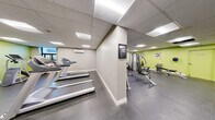 This fitness center offers a thoughtfully organized, single-floor layout that accommodates a wide range of exercise activities. The space is divided into distinct workout zones, each designed to support specific training needs. The cardio area features multiple treadmills and a stationary recumbent bike, arranged to allow comfortable spacing between users. A wall-mounted flat-screen TV overlooks the cardio zone, enhancing the workout experience. The strength training section is equipped with a multi-station cable machine, a dumbbell rack with a full range of free weights, an adjustable workout bench, and a functional trainer, providing comprehensive options for resistance training. The flooring throughout is durable rubber gym matting, ideal for high-impact activities and equipment use. Walls are finished in a combination of neutral gray and vibrant lime green accents, creating an energetic and motivating atmosphere. Recessed panel lighting in the drop ceiling ensures even, bright illumination across the entire space. A wall-mounted mirror extends along one side, visually expanding the room. Additional amenities include a wall-mounted water fountain, a sanitizing wipe dispenser, an air conditioning unit, and posted workout guides. Natural light is introduced through windows along the perimeter.