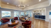 This well-appointed single-floor community space blends contemporary design with functional elegance. The expansive main lounge area commands attention with soaring ceilings adorned with a decorative textured finish and a striking circular chandelier as its centerpiece. Large glass-paned doors flood the space with natural light and provide views of the outdoor amenity area, while rich wood-look flooring runs throughout, anchoring the warm, inviting atmosphere.

The open-concept kitchen is a standout feature, showcasing crisp grey shaker cabinetry, white quartz countertops, a bold navy blue tiled island with gold-accented bar stools, and stainless steel appliances. Globe pendant lights hang above the island, adding a modern touch. A second kitchen or kitchenette area mirrors this aesthetic with patterned tile backsplash and ample counter space.

Adjacent workspaces include a welcoming reception desk and private offices accessible through elegant glass French doors, offering a professional yet approachable environment. A dedicated conference room features a large oval dining table with seating for six and an oversized window overlooking the outdoor area. Decorative gold-framed open shelving units against a deep navy accent wall add a sophisticated, curated finishing touch throughout the space.