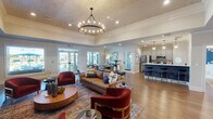 This well-appointed single-floor community space blends contemporary design with functional elegance. The expansive main lounge area commands attention with soaring ceilings adorned with a decorative textured finish and a striking circular chandelier as its centerpiece. Large glass-paned doors flood the space with natural light and provide views of the outdoor amenity area, while rich wood-look flooring runs throughout, anchoring the warm, inviting atmosphere.

The open-concept kitchen is a standout feature, showcasing crisp grey shaker cabinetry, white quartz countertops, a bold navy blue tiled island with gold-accented bar stools, and stainless steel appliances. Globe pendant lights hang above the island, adding a modern touch. A second kitchen or kitchenette area mirrors this aesthetic with patterned tile backsplash and ample counter space.

Adjacent workspaces include a welcoming reception desk and private offices accessible through elegant glass French doors, offering a professional yet approachable environment. A dedicated conference room features a large oval dining table with seating for six and an oversized window overlooking the outdoor area. Decorative gold-framed open shelving units against a deep navy accent wall add a sophisticated, curated finishing touch throughout the space.