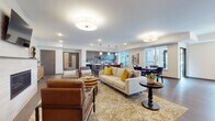 This thoughtfully designed single-floor community space offers an expansive, open-concept layout that seamlessly connects multiple functional zones. The main lounge area is anchored by a plush neutral sofa and leather accent chairs arranged atop a patterned area rug, centered around a sleek fireplace framed in light stone tile with a mounted television above. Warm hardwood-style flooring flows throughout, complementing the contemporary décor accented with mustard yellow cushions and earth-toned furnishings.

The full kitchen features dark cabinetry with a stone tile backsplash, a generous white-topped island with bar seating for several guests, and pendant lighting overhead. A dedicated catering or prep area with additional cabinetry and a compact refrigerator provides functional support for events and gatherings.

A built-in banquette dining nook offers intimate seating with a yellow upholstered bench, café-style tables, and framed black-and-white photography on textured wall panels. A separate dining area with a tray ceiling and large window frames lush outdoor greenery, filling the space with natural light.

Recessed lighting and drum-style flush-mount fixtures provide consistent illumination throughout. The overall ambiance is modern, welcoming, and versatile — well suited for both casual socializing and organized group activities.