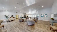 This well-appointed single-floor facility offers a thoughtfully designed layout encompassing multiple distinct gathering and lounge areas, all unified by warm wood-look flooring and a clean, contemporary aesthetic. The main hall is expansive and open, featuring soaring vaulted ceilings with shiplap detailing, ceiling fans, and recessed lighting that create a bright and welcoming atmosphere. Multiple seating arrangements — including midcentury-modern accent chairs and sofas — are distributed across the space to encourage both intimate conversation and larger group gatherings. A dedicated lounge area with teal upholstered chairs opens toward exterior doors, allowing natural light to filter in through large plantation-shuttered windows. Additional rooms include a comfortable sitting room with board-and-batten walling, a spacious entertainment room with vaulted ceilings and a wall-mounted television, and a well-equipped full kitchen outfitted with white shaker cabinetry, granite-style countertops, stainless steel appliances, a tiled backsplash, and a skylight overhead. The entrance foyer welcomes visitors with high ceilings, French doors, and tasteful décor. Throughout, the neutral white walls and natural light contribute to an airy, polished, and functional environment.
