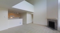 This two-story space is defined by its dramatic double-height ceilings in the main living area, creating an expansive, open atmosphere that immediately draws the eye upward. The upper level features a mezzanine-style overhang, adding architectural depth and visual interest to the layout. The living area is finished with neutral wall-to-wall carpeting, contributing to a clean, understated aesthetic throughout.

The kitchen is positioned adjacent to the living space and is equipped with light wood cabinetry, a built-in microwave, and a countertop peninsula that provides both workspace and a natural divide between the kitchen and living areas. Track lighting is installed above the kitchen, ensuring ample illumination over the functional zones.

Neutral, warm tones dominate the interior palette, from the cream-colored walls to the light carpet underfoot, lending the space a cohesive and calm ambiance. A doorway visible from the main living area leads to additional rooms beyond, suggesting a practical floor plan. Overall, the combination of soaring ceilings, an open-concept design, and consistent neutral finishes creates a versatile, light-filled environment.