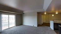 This single-floor residential space offers a clean, functional layout designed for comfortable everyday living. The main living area is generously sized, finished with soft wall-to-wall carpeting that adds warmth underfoot. A distinctive tray ceiling adds architectural interest overhead, creating a subtle sense of depth and dimension to the room. Neutral gray-toned walls provide a versatile backdrop, well-suited for a variety of décor styles. A sliding glass patio door along one wall invites natural light into the space and provides direct access to an outdoor balcony or patio area, enhancing the connection between interior and exterior living. A baseboard heating unit runs along the base of the interior wall, ensuring efficient climate control throughout the space. The living area flows naturally into a connected entryway or hallway, which features darker hardwood-style flooring, offering a clean visual contrast. This secondary area is illuminated by a ceiling-mounted light fixture and leads to a paneled entry door, providing a welcoming transition into the home. The overall ambiance of the space is understated and move-in ready, characterized by its open feel, thoughtful layout, and practical finishes throughout.