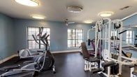 Located at 10900 E Taylor Rd in Gulfport, Mississippi, this single-floor fitness center is a well-equipped workout facility designed to support a comprehensive exercise routine. The space is finished with durable dark rubber flooring throughout, providing a safe and functional surface for a wide range of activities.

The room is outfitted with a variety of cardio machines, including multiple elliptical trainers, treadmills, and a stationary bike, offering ample options for aerobic workouts. Strength training is supported by a multi-station cable machine, a weight bench, and free weights, catering to users of varying fitness levels. A step platform and additional small equipment are also available for floor-based exercises.

Large wall-mounted mirrors line one side of the room, visually expanding the space and allowing users to monitor their form during workouts. Multiple windows allow natural light to filter in and offer views of an outdoor pool area, creating an energizing atmosphere. A ceiling fan provides additional air circulation, complementing the overhead flush-mount lighting that keeps the space bright and well-lit.

The walls are painted in a calming light blue tone, and a Coca-Cola vending machine is conveniently positioned near the entrance. A wall-mounted display and sanitation station are also present, reflecting attention to cleanliness and usability. The facility is accessible via a standard interior door.