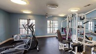 Located at 10900 E Taylor Rd in Gulfport, Mississippi, this single-floor fitness center is a well-equipped workout facility designed to support a comprehensive exercise routine. The space is finished with durable dark rubber flooring throughout, providing a safe and functional surface for a wide range of activities.

The room is outfitted with a variety of cardio machines, including multiple elliptical trainers, treadmills, and a stationary bike, offering ample options for aerobic workouts. Strength training is supported by a multi-station cable machine, a weight bench, and free weights, catering to users of varying fitness levels. A step platform and additional small equipment are also available for floor-based exercises.

Large wall-mounted mirrors line one side of the room, visually expanding the space and allowing users to monitor their form during workouts. Multiple windows allow natural light to filter in and offer views of an outdoor pool area, creating an energizing atmosphere. A ceiling fan provides additional air circulation, complementing the overhead flush-mount lighting that keeps the space bright and well-lit.

The walls are painted in a calming light blue tone, and a Coca-Cola vending machine is conveniently positioned near the entrance. A wall-mounted display and sanitation station are also present, reflecting attention to cleanliness and usability. The facility is accessible via a standard interior door.