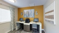 This single-floor business center is thoughtfully designed to provide a functional and visually engaging workspace. The room's most striking feature is a bold amber accent wall that serves as a dynamic focal point, adorned with a large abstract cityscape painting flanked by decorative "@" and "&" symbols, adding a modern, urban touch to the space.

Two granite-topped workstations are positioned side by side along the accent wall, each equipped with a desktop computer and accompanied by sleek, contemporary grey chairs. A shared multifunction printer sits centrally between the two stations, ensuring convenient access for all users. The workstations offer ample surface area for productive use, making this an ideal setup for residents or guests needing to handle business tasks.

The flooring features warm wood-tone planks that complement the room's color palette, while patterned curtains on the side window introduce a soft, layered texture to the overall design. Overhead, flush-mounted lighting provides consistent, even illumination throughout the room. The space strikes a balance between professional utility and inviting aesthetics, making it a comfortable and efficient environment for work, printing, and connectivity.