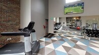 Located in Silverdale, WA, this single-floor fitness center offers a thoughtfully designed workout environment that balances open space with defined functional zones. The facility is characterized by soaring vaulted ceilings equipped with skylights and recessed lighting, flooding the interior with natural and artificial light throughout the day. High clerestory windows further enhance brightness while maintaining an energetic atmosphere.

The main exercise floor is covered in a bold geometric tile pattern in blue, gray, and white tones, creating a visually dynamic foundation. A dedicated functional training zone features a dark sport-turf mat printed with track-style lane markings, anchored by a central multi-use cable and strength rig. This area accommodates battle ropes, medicine balls, and suspension training accessories.

Surrounding the functional zone, a comprehensive selection of Life Fitness selectorized strength machines is arranged to allow easy circulation. A cardio section houses treadmills and steppers positioned near wall-mounted flat-screen televisions. Full-length mirrors expand the sense of space and aid in form monitoring. A glass-partitioned area adjacent to the main floor provides visual connectivity while defining a separate training enclosure.

An exposed brick accent wall with a large window frames views of surrounding greenery, adding warmth and texture to the otherwise contemporary interior. A glass-railed staircase connects to an upper level, completing this well-equipped facility.