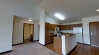 This single-floor space is designed with functionality and comfort in mind. The kitchen serves as a central feature, showcasing warm medium-toned wood cabinetry that lines the walls and provides generous storage. A peninsula with a granite-style countertop extends into the living area, creating a natural breakfast bar that separates the kitchen from the rest of the space while maintaining an open, connected feel. The kitchen is equipped with white appliances, including a range, microwave, refrigerator, and a double-basin sink built into the peninsula. Flooring transitions from hardwood in the kitchen to carpet in the adjacent living area, adding textural variety and defining each zone. The ceilings are elevated, contributing to a sense of openness and airiness throughout. Flush-mounted lighting fixtures provide consistent, even illumination across the space. A hallway visible from the kitchen leads to a full bathroom, which features a bathtub, toilet, and bright overhead lighting. The overall color palette is neutral and understated, with white walls and trim complementing the wood tones of the cabinetry, creating a clean and welcoming environment throughout.