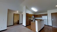 This single-floor space is designed with functionality and comfort in mind. The kitchen serves as a central feature, showcasing warm medium-toned wood cabinetry that lines the walls and provides generous storage. A peninsula with a granite-style countertop extends into the living area, creating a natural breakfast bar that separates the kitchen from the rest of the space while maintaining an open, connected feel. The kitchen is equipped with white appliances, including a range, microwave, refrigerator, and a double-basin sink built into the peninsula. Flooring transitions from hardwood in the kitchen to carpet in the adjacent living area, adding textural variety and defining each zone. The ceilings are elevated, contributing to a sense of openness and airiness throughout. Flush-mounted lighting fixtures provide consistent, even illumination across the space. A hallway visible from the kitchen leads to a full bathroom, which features a bathtub, toilet, and bright overhead lighting. The overall color palette is neutral and understated, with white walls and trim complementing the wood tones of the cabinetry, creating a clean and welcoming environment throughout.