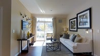 This single-floor residence showcases a well-appointed living area designed with both comfort and style in mind. The space is anchored by rich hardwood flooring that runs the length of the room, complementing the warm, neutral tones of the walls. A large sliding glass door serves as a focal point, flooding the interior with abundant natural light while providing direct access to a private balcony with views of the surrounding neighborhood. Elegant sheer curtains frame the door, adding softness to the space.

The living area is thoughtfully arranged with a plush white sofa accented by decorative pillows in grey and earth tones, paired with a geometric metal-framed glass coffee table that adds a contemporary touch. A wall-mounted flat-screen television is positioned across from the seating area, accompanied by a sleek console table adorned with decorative objects and floral arrangements in muted yellow and green tones. A framed abstract artwork on the adjacent wall contributes an artistic element to the overall aesthetic. The space conveys a clean, modern ambiance that balances functionality with refined residential design.