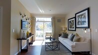 This single-floor residence showcases a well-appointed living area designed with both comfort and style in mind. The space is anchored by rich hardwood flooring that runs the length of the room, complementing the warm, neutral tones of the walls. A large sliding glass door serves as a focal point, flooding the interior with abundant natural light while providing direct access to a private balcony with views of the surrounding neighborhood. Elegant sheer curtains frame the door, adding softness to the space.

The living area is thoughtfully arranged with a plush white sofa accented by decorative pillows in grey and earth tones, paired with a geometric metal-framed glass coffee table that adds a contemporary touch. A wall-mounted flat-screen television is positioned across from the seating area, accompanied by a sleek console table adorned with decorative objects and floral arrangements in muted yellow and green tones. A framed abstract artwork on the adjacent wall contributes an artistic element to the overall aesthetic. The space conveys a clean, modern ambiance that balances functionality with refined residential design.