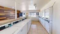 Located at 1950 North 43rd Avenue in Phoenix, Arizona, this single-floor community laundry facility offers a well-organized and functional space designed to accommodate high-volume laundry needs. The layout is divided into two connected areas, together providing a generous amount of floor space for comfortable use by multiple patrons simultaneously.

The main laundry area is lined with rows of coin-operated top-load washing machines along one wall and a bank of stacked front-load dryers along another, offering substantial capacity for residents. Laminate folding counters run alongside the machines, providing convenient surfaces for sorting and folding laundry. The space is finished with light-toned ceramic tile flooring throughout, which is both durable and easy to maintain.

A standout design feature is a full accent wall clad in reclaimed wood planks in varying natural tones — from warm amber and honey to deep walnut — adding a rustic, visually striking contrast to the otherwise utilitarian environment. Natural light filters in through barred windows, complementing the overhead fluorescent lighting to keep the space bright and well-lit. The textured white ceilings and clean white walls maintain a crisp, practical aesthetic. Posted informational signage ensures users have clear guidance on machine operation and facility rules.