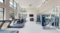 Located at 4545 South Blvd in Virginia Beach, Virginia, this single-floor fitness center is designed to deliver a premium workout experience in a bright, modern environment. The space is defined by its dramatic double-height ceilings, which lend an open, airy atmosphere throughout. Two tiers of large, black-framed windows wrap the perimeter, flooding the interior with natural light and offering views of the surrounding greenery and community.

The fitness center is equipped with a comprehensive selection of cardio machines, including multiple treadmills and elliptical trainers, all arranged along the windowed walls to maximize the sense of openness. A dedicated strength training zone features a multi-station cable machine system, weight benches, and a full rack of dumbbells, complemented by full-length mirrors that enhance both the visual depth of the space and workout functionality. Additional specialized equipment, such as a leg press machine and an air bike, rounds out the facility's offerings.

Light-toned luxury vinyl flooring runs throughout, contributing to the clean, contemporary aesthetic. Recessed lighting and a ceiling fan ensure comfort during all seasons. A wall-mounted flat-screen television adds entertainment to cardio sessions. Built-in cubbies provide convenient storage, and a dedicated entry door with posted fitness center guidelines completes this well-appointed facility.