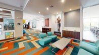 This well-appointed fitness center in Davie, Florida, offers a comprehensive and thoughtfully designed workout environment across a single floor. Guests are welcomed by a striking lobby entrance featuring dramatic pendant lighting, floor-to-ceiling glass walls, and a sleek staircase with glass railings — setting an upscale tone from the moment of arrival.

The main corridor is immediately eye-catching, with a bold, geometric chevron-patterned carpet in teal, orange, and cream tones that carries throughout the facility. Glass-partitioned rooms line the hallway, lending an open, transparent feel to the space. A stylish lounge area provides teal seating, a low coffee table, and athletic-themed wall art, offering a comfortable space to relax before or after a workout.

The cardio zone is equipped with a row of modern treadmills positioned alongside large windows overlooking lush outdoor palm trees, as well as air bikes and recumbent cycles complemented by vibrant teal accent walls and contemporary artwork. The strength training area is generously outfitted with a full rack of free weights, multiple cable and resistance machines, and wall-to-wall mirrors that amplify the sense of space. Wall-mounted televisions are positioned throughout for entertainment. Functional training accessories, including stability balls and balance trainers, round out the facility's diverse offerings.