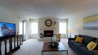 This single-floor residence offers a thoughtfully designed living space centered around a striking electric fireplace set into a clean, white-painted accent wall. A decorative round mirror mounted above the fireplace adds a stylish focal point to the room, complementing the understated, contemporary aesthetic throughout.

The living area is finished with wall-to-wall neutral carpeting that adds warmth underfoot, while the textured ceiling contributes subtle architectural character. Natural light fills the space generously through multiple windows dressed with white curtains and horizontal blinds, creating a bright and airy atmosphere. A sliding glass door on one side of the room provides additional light and access to an outdoor area.

The room is well-proportioned, offering ample space for comfortable seating arrangements. A dark sofa and an accent chair with mustard yellow cushions provide stylish yet practical furnishings, anchored by a wood-and-metal coffee table at the center. The overall color palette is composed of soft whites, warm neutrals, and muted grays, lending the space a calm and cohesive feel.

The layout flows naturally, making the living area feel open and functional. The combination of the fireplace, abundant natural light, and carefully chosen furnishings creates an inviting environment well-suited for both relaxed everyday living and casual entertaining.