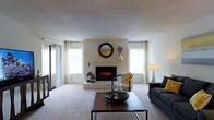 This single-floor residence offers a thoughtfully designed living space centered around a striking electric fireplace set into a clean, white-painted accent wall. A decorative round mirror mounted above the fireplace adds a stylish focal point to the room, complementing the understated, contemporary aesthetic throughout.

The living area is finished with wall-to-wall neutral carpeting that adds warmth underfoot, while the textured ceiling contributes subtle architectural character. Natural light fills the space generously through multiple windows dressed with white curtains and horizontal blinds, creating a bright and airy atmosphere. A sliding glass door on one side of the room provides additional light and access to an outdoor area.

The room is well-proportioned, offering ample space for comfortable seating arrangements. A dark sofa and an accent chair with mustard yellow cushions provide stylish yet practical furnishings, anchored by a wood-and-metal coffee table at the center. The overall color palette is composed of soft whites, warm neutrals, and muted grays, lending the space a calm and cohesive feel.

The layout flows naturally, making the living area feel open and functional. The combination of the fireplace, abundant natural light, and carefully chosen furnishings creates an inviting environment well-suited for both relaxed everyday living and casual entertaining.