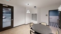 This single-floor commercial space combines contemporary design with practical functionality across its well-appointed layout. The main common area serves as the heart of the suite, anchored by a round dark matte table surrounded by cane-back chairs in black frames. Elegant pleated pendant lights hang overhead, casting warm, focused light against crisp white walls and light herringbone-patterned flooring that lends a refined, modern character to the space. Built-in grey lockers are recessed into the wall, providing secure personal storage. A textured grey accent wall with a built-in counter and cabinetry adds depth and a sophisticated finishing touch to the area. A corridor connects the main space to additional zones, featuring clean white walls, dark-framed glass doors, and smart thermostat controls. The hallway opens toward what appears to be an elevator access point and additional seating with warm, padded banquette benches alongside a textured feature wall. The restroom is designed with accessibility in mind, featuring stainless steel grab bars, recessed fixtures, and a striking combination of glossy white subway tile wainscoting set against a rich teal upper wall. A bold geometric patterned floor in navy, teal, and white completes the restroom's polished, detail-oriented aesthetic. Overall, the space presents a sleek, professional environment suited for modern commercial use.