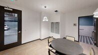 This single-floor commercial space combines contemporary design with practical functionality across its well-appointed layout. The main common area serves as the heart of the suite, anchored by a round dark matte table surrounded by cane-back chairs in black frames. Elegant pleated pendant lights hang overhead, casting warm, focused light against crisp white walls and light herringbone-patterned flooring that lends a refined, modern character to the space. Built-in grey lockers are recessed into the wall, providing secure personal storage. A textured grey accent wall with a built-in counter and cabinetry adds depth and a sophisticated finishing touch to the area. A corridor connects the main space to additional zones, featuring clean white walls, dark-framed glass doors, and smart thermostat controls. The hallway opens toward what appears to be an elevator access point and additional seating with warm, padded banquette benches alongside a textured feature wall. The restroom is designed with accessibility in mind, featuring stainless steel grab bars, recessed fixtures, and a striking combination of glossy white subway tile wainscoting set against a rich teal upper wall. A bold geometric patterned floor in navy, teal, and white completes the restroom's polished, detail-oriented aesthetic. Overall, the space presents a sleek, professional environment suited for modern commercial use.
