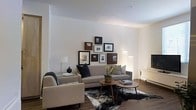 This single-floor space showcases a thoughtfully designed interior that balances modern aesthetics with a warm, inviting atmosphere. The living area serves as the centerpiece, featuring a neutral-toned sofa and accent chair arrangement anchored by a striking cowhide area rug. A pair of mid-century modern coffee tables with tapered legs adds a classic yet contemporary touch to the layout.

One of the standout features is an artfully arranged gallery wall, displaying a diverse collection of framed artwork in varying sizes and finishes, creating a visually dynamic focal point. Soft, layered lighting from a floor lamp and recessed ceiling fixtures bathes the room in a warm glow, complementing the natural tones throughout.

The space also includes a sleek fireplace set into a clean, minimalist surround, adding both function and architectural interest. High ceilings enhance the sense of openness, while white walls provide a crisp backdrop that allows the furnishings and décor to stand out. Hardwood flooring runs throughout, contributing warmth and continuity. Overall, this space presents a refined, contemporary environment well-suited for comfortable everyday living and stylish entertaining.
