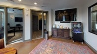 This thoughtfully designed single-floor space blends comfort and professionalism seamlessly. The main lounge area serves as the heart of the space, featuring rich hardwood flooring, warm gray walls, and an abundance of natural light streaming through multiple double-hung windows with dark-trimmed frames. A classic tufted leather sofa anchors the seating area, accompanied by worn leather armchairs and a rustic wooden coffee table, creating an inviting and relaxed atmosphere. A series of large mirrors along one wall expand the visual depth of the room, while framed artwork and wall-mounted sconces add refined touches throughout.

Adjacent to the lounge, a dining or meeting area offers round wooden tables and upholstered chairs set atop a vibrant patterned area rug, ideal for small gatherings or consultations. A large flat-screen television is mounted above a dark wood credenza stocked with refreshments and beverages, providing a convenient hospitality station for guests. Sliding doors lead to additional rooms beyond, suggesting a functional back-of-house area.

A private workstation room features carpet tile flooring, a granite-topped desk, and a computer setup beside a window with horizontal blinds, offering a quiet and focused environment. Throughout the space, recessed lighting and track lighting complement the natural light, ensuring a bright and welcoming ambiance across all areas.