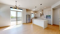 This contemporary single-floor residence is thoughtfully designed with a clean, modern aesthetic throughout. The open-concept layout seamlessly connects the kitchen and living areas, creating a bright and airy atmosphere enhanced by abundant natural light streaming through a series of clerestory and full-height windows.

The kitchen is a standout feature, showcasing crisp white shaker cabinetry with matte black hardware, a striking marble-style tile backsplash, and a generous central island topped with a smooth white quartz countertop. Stainless steel appliances — including a French door refrigerator, wall oven, and built-in microwave — complete the well-appointed cooking space. A stainless steel range hood adds an industrial touch, while stylish pendant lights hang above the island, complemented by recessed ceiling lighting throughout.

Light-toned hardwood flooring flows continuously across the entire residence, lending warmth and cohesion to each room. The bedrooms are well-proportioned, featuring ceiling fans, horizontal clerestory windows that frame views of surrounding greenery, and clean white walls that offer a versatile, neutral canvas. One room opens directly onto a spacious outdoor terrace through sliding glass doors, providing an inviting extension of the living space with open views of the surrounding landscape and treetops.