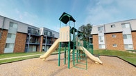 This outdoor playground area serves as a central amenity within a residential community. The focal point is a well-maintained, multi-level play structure finished in a green and beige color scheme. The structure features two wide slides extending from an elevated central tower, which is topped with a dark green roof panel. Sturdy green metal framing supports the platform, while arched ladder entries on either side provide easy access for children of varying ages. The play structure is designed to encourage active play and social interaction among younger residents.

The ground beneath the structure is covered in rubberized mulch surfacing, providing a safe and impact-absorbing material around the equipment. The surrounding area features a well-kept lawn with patches of green grass, adding a natural backdrop to the space. Mature trees line the perimeter, offering partial shade on sunny days.

The playground is nestled between multi-story residential buildings constructed of brick and siding, with balconies overlooking the open area. The setting creates a sheltered, community-oriented atmosphere while maintaining an open-air feel under an open sky. The combination of durable equipment, safe surfacing, and accessible design makes this playground a practical and inviting outdoor space for residents.