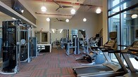 This two-story fitness center offers a comprehensive workout environment designed to accommodate a wide range of exercise needs. The space is outfitted with an extensive selection of modern equipment, including ellipticals, treadmills, and multi-station strength machines from Matrix and other leading brands, arranged to allow comfortable movement between stations.

The interior features generous ceiling heights that contribute to an open, airy atmosphere. Pendant lighting fixtures are suspended throughout, providing bright, even illumination across the floor, while ceiling fans promote air circulation during workouts. A bold patterned carpet in dark tones with red accents lines the floor, adding a dynamic visual element to the space.

Large mirrors are mounted along the walls, enhancing the sense of depth and allowing users to monitor their form during exercise. A staircase connects the two floors, suggesting additional workout or amenity space above. The exposed red accent piping along the ceiling adds an industrial design touch that contrasts effectively with the modern equipment and clean layout, creating a motivating and functional fitness environment.