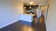 This single-floor space showcases an open-concept layout that seamlessly integrates the kitchen and living areas. The kitchen is equipped with a full suite of stainless steel appliances, including a refrigerator, gas range, and built-in microwave. Dark espresso-toned cabinetry extends to the upper and lower sections of the kitchen walls, complemented by a subway tile backsplash that adds a clean, contemporary aesthetic. A peninsula with light-toned stone countertops serves as both a food preparation area and a breakfast bar, providing additional seating and a natural division between the kitchen and the open living space. Pendant lighting hangs above the peninsula and kitchen work areas, enhancing both function and ambiance. Rich, dark hardwood flooring spans the entirety of the space, providing a striking contrast against the neutral-toned walls. The generous open floor plan allows for flexible furniture arrangement in the living area. A doorway visible in the background suggests additional rooms beyond, contributing to the overall sense of a well-organized and functional layout.