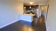 This single-floor space showcases an open-concept layout that seamlessly integrates the kitchen and living areas. The kitchen is equipped with a full suite of stainless steel appliances, including a refrigerator, gas range, and built-in microwave. Dark espresso-toned cabinetry extends to the upper and lower sections of the kitchen walls, complemented by a subway tile backsplash that adds a clean, contemporary aesthetic. A peninsula with light-toned stone countertops serves as both a food preparation area and a breakfast bar, providing additional seating and a natural division between the kitchen and the open living space. Pendant lighting hangs above the peninsula and kitchen work areas, enhancing both function and ambiance. Rich, dark hardwood flooring spans the entirety of the space, providing a striking contrast against the neutral-toned walls. The generous open floor plan allows for flexible furniture arrangement in the living area. A doorway visible in the background suggests additional rooms beyond, contributing to the overall sense of a well-organized and functional layout.