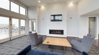 This single-floor space combines contemporary design with functional amenities in an open, airy layout. The lounge area is a standout feature, characterized by dramatically high ceilings and an expansive wall of floor-to-ceiling windows that frame sweeping waterfront views. Natural light floods the interior, creating a bright and inviting atmosphere. A statement fireplace clad in white stacked stone anchors one wall, topped by a mounted flat-screen television, with comfortable seating arranged around a clean-lined wood coffee table to encourage gathering.

The kitchen area is outfitted with rich, dark-stained cabinetry paired with granite countertops, stainless steel appliances including a full-size refrigerator, dishwasher, and microwave, and a deep double sink. Wood-look flooring runs throughout the kitchen and dining zone, where a dining table with wooden chairs provides ample seating. A frosted glass partition offers a subtle division between spaces while maintaining an open feel. Recessed lighting throughout ensures consistent illumination, and an adjoining outdoor balcony extends the waterfront experience beyond the interior.