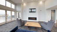 This single-floor space combines contemporary design with functional amenities in an open, airy layout. The lounge area is a standout feature, characterized by dramatically high ceilings and an expansive wall of floor-to-ceiling windows that frame sweeping waterfront views. Natural light floods the interior, creating a bright and inviting atmosphere. A statement fireplace clad in white stacked stone anchors one wall, topped by a mounted flat-screen television, with comfortable seating arranged around a clean-lined wood coffee table to encourage gathering.

The kitchen area is outfitted with rich, dark-stained cabinetry paired with granite countertops, stainless steel appliances including a full-size refrigerator, dishwasher, and microwave, and a deep double sink. Wood-look flooring runs throughout the kitchen and dining zone, where a dining table with wooden chairs provides ample seating. A frosted glass partition offers a subtle division between spaces while maintaining an open feel. Recessed lighting throughout ensures consistent illumination, and an adjoining outdoor balcony extends the waterfront experience beyond the interior.