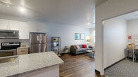 This well-designed 3-bedroom, 2-bathroom unit offers a thoughtful single-floor layout that maximizes functionality. The open-concept living and kitchen area serves as the heart of the home, featuring rich wood-style flooring that flows seamlessly throughout the main living spaces. The kitchen is equipped with stainless steel appliances, including a full-size refrigerator, electric range, microwave, and dishwasher. Granite countertops extend along ample cabinet space as well as a peninsula breakfast bar with seating for two, making it ideal for casual dining and entertaining. White upper cabinetry and track lighting complete the kitchen's clean, modern aesthetic. The living room is bright and inviting, anchored by a large window that fills the space with natural light. A striking teal accent wall adds a contemporary pop of color. Each of the three bedrooms features plush carpet flooring and a generously sized closet with built-in shelving. The primary bedroom offers a comfortable, private retreat. Both bathrooms are finished with granite vanity countertops, wood-style flooring, and glass-door walk-in showers. Clean lines, neutral tones, and modern finishes throughout give this unit a cohesive and welcoming atmosphere.