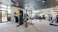 This expansive fitness center offers a comprehensively equipped workout environment designed for a wide range of exercise routines. The space is divided into two distinct zones: a main gym floor and a separate group fitness studio, each thoughtfully appointed with purpose-specific flooring — geometric-patterned carpet tile in the main gym area and smooth hardwood in the studio.

The main gym floor is outfitted with an extensive selection of equipment, including treadmills, ellipticals, a stair climber, cable machines, multi-functional strength stations, selectorized machines, a full dumbbell rack, and kettlebells. Full-length mirrors line the walls, amplifying the sense of space and allowing users to monitor their form. Overhead, dark-painted ceilings with ceiling fans, recessed lighting, and suspended linear pendant fixtures lend a modern, polished atmosphere.

The adjoining studio features a large, open hardwood floor ideal for yoga, stretching, cycling, or group classes. It is furnished with spin bikes, stability balls, medicine balls, and resistance accessories. Floor-to-ceiling black-framed glass walls and oversized windows provide abundant natural light and outdoor views, creating an energizing environment. A retractable glass garage-style door connects the studio to the main gym, offering flexible, open-concept use of the overall space.