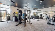 This expansive fitness center offers a comprehensively equipped workout environment designed for a wide range of exercise routines. The space is divided into two distinct zones: a main gym floor and a separate group fitness studio, each thoughtfully appointed with purpose-specific flooring — geometric-patterned carpet tile in the main gym area and smooth hardwood in the studio.

The main gym floor is outfitted with an extensive selection of equipment, including treadmills, ellipticals, a stair climber, cable machines, multi-functional strength stations, selectorized machines, a full dumbbell rack, and kettlebells. Full-length mirrors line the walls, amplifying the sense of space and allowing users to monitor their form. Overhead, dark-painted ceilings with ceiling fans, recessed lighting, and suspended linear pendant fixtures lend a modern, polished atmosphere.

The adjoining studio features a large, open hardwood floor ideal for yoga, stretching, cycling, or group classes. It is furnished with spin bikes, stability balls, medicine balls, and resistance accessories. Floor-to-ceiling black-framed glass walls and oversized windows provide abundant natural light and outdoor views, creating an energizing environment. A retractable glass garage-style door connects the studio to the main gym, offering flexible, open-concept use of the overall space.