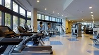 This modern fitness center offers a spacious, single-floor layout designed to accommodate a comprehensive workout experience. The facility is anchored by an impressive wall of floor-to-ceiling windows that floods the space with natural light and provides open views of the surrounding greenery, creating an energizing and welcoming atmosphere.

The cardio zone is prominently featured along the windowed perimeter, housing a well-curated lineup of treadmills, elliptical trainers, and stair climbers from reputable brands including Matrix and Cybex. The equipment is arranged in an orderly fashion, allowing for comfortable spacing between machines and ease of movement throughout the floor.

Complementing the cardio area, a dedicated strength training section features multi-functional cable and weight machines positioned along the far end of the facility. The clean, two-tone blue and white vinyl flooring adds a professional aesthetic while providing a durable, easy-to-maintain surface.

Recessed ceiling lighting works in tandem with the abundant natural light to ensure the space is consistently bright and well-illuminated at all hours. The neutral color palette of whites and grays, combined with the open layout and high ceilings, contributes to a clean, contemporary ambiance that is both functional and visually appealing.