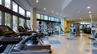 This modern fitness center offers a spacious, single-floor layout designed to accommodate a comprehensive workout experience. The facility is anchored by an impressive wall of floor-to-ceiling windows that floods the space with natural light and provides open views of the surrounding greenery, creating an energizing and welcoming atmosphere.

The cardio zone is prominently featured along the windowed perimeter, housing a well-curated lineup of treadmills, elliptical trainers, and stair climbers from reputable brands including Matrix and Cybex. The equipment is arranged in an orderly fashion, allowing for comfortable spacing between machines and ease of movement throughout the floor.

Complementing the cardio area, a dedicated strength training section features multi-functional cable and weight machines positioned along the far end of the facility. The clean, two-tone blue and white vinyl flooring adds a professional aesthetic while providing a durable, easy-to-maintain surface.

Recessed ceiling lighting works in tandem with the abundant natural light to ensure the space is consistently bright and well-illuminated at all hours. The neutral color palette of whites and grays, combined with the open layout and high ceilings, contributes to a clean, contemporary ambiance that is both functional and visually appealing.