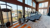 Located at 2019 Midway Rd in Lewisville, TX, this fitness center is a comprehensively outfitted facility designed to support a wide range of workout styles and fitness goals. The space is organized into distinct functional zones that flow seamlessly into one another, creating an efficient and motivating environment for exercise.

The cardio area is positioned along a dramatic wall of floor-to-ceiling windows that rise to soaring double-height ceilings, offering unobstructed views of an outdoor pool and landscaped surroundings. Rows of treadmills, ellipticals, and a stair climber are arranged to take full advantage of this natural light and outdoor scenery. The patterned carpet features bold orange stripe accents that add visual energy to the space.

A dedicated strength training zone features a multi-station cable machine, weight benches, and a well-stocked dumbbell rack reflected in full-length mirrors that amplify the sense of space. Adjoining this area is a studio-style room with hardwood-style flooring, spinning bikes, additional free weights, medicine balls, and a large display screen for guided workouts. Plyosoft boxes and functional training tools, including a battle rope station and a heavy bag, round out the facility's offerings, ensuring a truly versatile training environment throughout.