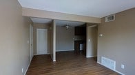 This single-floor space features an open-concept layout that seamlessly connects the living area to the kitchen, creating a cohesive and functional flow throughout. Rich, dark hardwood flooring extends across the entire space, providing a warm and grounded aesthetic that ties the rooms together.

The living area is generously proportioned, with neutral taupe-toned walls and a textured ceiling that adds subtle dimension overhead. A ceiling fan is centrally positioned in the adjacent room, offering both practical ventilation and a clean, classic look. A floor vent ensures efficient climate control throughout the space.

The kitchen is partially open to the living area, defined by a structural beam overhead that frames the transition between spaces. Dark wood cabinetry lines the kitchen walls, offering ample storage, while the included appliances add immediate functionality. The overall kitchen design is practical and straightforward.

Near the entryway, a set of bifold closet doors provides convenient storage space, complementing the home's practical layout. The front door is positioned for easy access. The neutral color palette throughout — from the walls to the trim — creates a clean, move-in-ready canvas well-suited for a variety of interior styles.