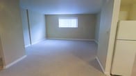 This single-floor space presents a clean, straightforward layout designed for functional living. The main room is generously proportioned, offering an open and airy feel with ample room to accommodate a variety of furniture arrangements. The flooring is covered in plush, light-toned wall-to-wall carpeting, providing a soft and comfortable underfoot experience throughout the space. The walls are finished in a warm, neutral beige tone that contributes to a calm and understated ambiance. A centrally positioned window fitted with vertical blinds allows for adjustable natural light, brightening the interior while maintaining privacy. White baseboards frame the perimeter of the room, adding a crisp contrast to the neutral wall color. The overall design is minimal and move-in ready, offering a blank canvas well-suited for personalization. The single-level layout ensures easy accessibility and seamless flow throughout the space.