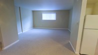 This single-floor space presents a clean, straightforward layout designed for functional living. The main room is generously proportioned, offering an open and airy feel with ample room to accommodate a variety of furniture arrangements. The flooring is covered in plush, light-toned wall-to-wall carpeting, providing a soft and comfortable underfoot experience throughout the space. The walls are finished in a warm, neutral beige tone that contributes to a calm and understated ambiance. A centrally positioned window fitted with vertical blinds allows for adjustable natural light, brightening the interior while maintaining privacy. White baseboards frame the perimeter of the room, adding a crisp contrast to the neutral wall color. The overall design is minimal and move-in ready, offering a blank canvas well-suited for personalization. The single-level layout ensures easy accessibility and seamless flow throughout the space.