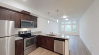 This single-floor residence features an open-concept layout that seamlessly connects the kitchen and living area, creating a bright and functional living environment. The kitchen is outfitted with rich dark-wood cabinetry that provides ample storage, complemented by light granite countertops with subtle veining. Stainless steel appliances, including a range, over-the-range microwave, and dishwasher, add a polished, modern aesthetic. A peninsula with an undermount sink extends the counter space and serves as a natural divider between the kitchen and the adjoining living area. Pendant lighting hangs above the peninsula, offering focused task lighting while enhancing the overall design. The flooring throughout is a light-toned hardwood, contributing to the airy feel of the space. Large windows in the living area flood the space with natural light and frame views of mature surrounding trees, bringing a sense of the outdoors inside. The white walls and flush ceiling fixtures further enhance the brightness of the space, giving it a clean, contemporary feel throughout.